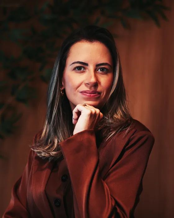 Dra. Raphaela Meyge com a mão no queixo, diante de um painel de madeira